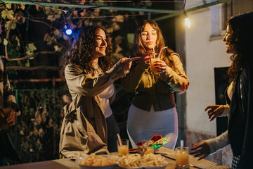 A group of friends gathers outdoors at night, enjoying drinks, snacks, and joyous company during a birthday celebration. Decorative lighting adds a festive and warm ambiance to the occasion.