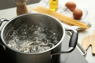 Cooking pot with boiling water and stove on white table, closeup