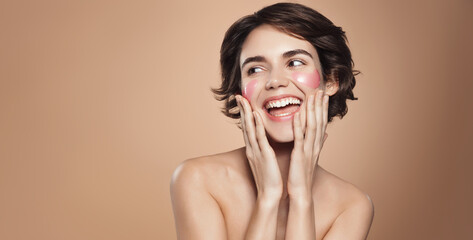 Portrait of beautiful, happy young woman with short hair, laughing, uses under eye patches to get this glazy donut skin, flawless look after skincare treatment, looks aside, brown background © Liubov Levytska