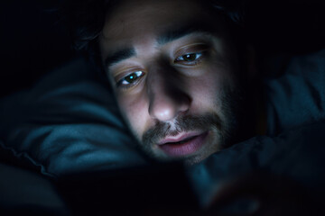 A man lies in bed, gazing intently at his smartphone screen as he struggles with insomnia in a darkened room