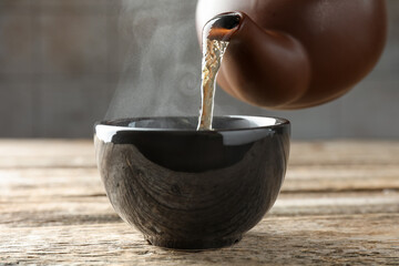 Pouring aromatic tea from teapot into cup at wooden table against grey background, closeup