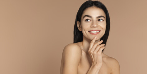 Glazy donut skin. Beautiful young woman with radiant flawless face, smiling and looking dreamy at empty space, touching her chin, standing over brown background © Liubov Levytska