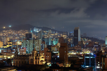 Fototapeta premium Breathtaking night view of Medellín, Medellin, Antioquia, Colombia, with illuminated skyscrapers and historic architecture. The city glows against the Andean mountains, creating a urban landscape.