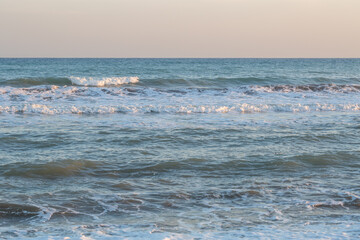 Crystal blue waves roll gently toward the shore under a pastel sky at sunset. The rhythmic movement of the sea creates a serene and calming atmosphere perfect for relaxation and reflection.