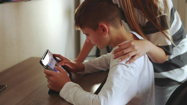 Teenage sister providing caring support while younger brother engages with tablet, showcasing familial love and digital interaction within home setting