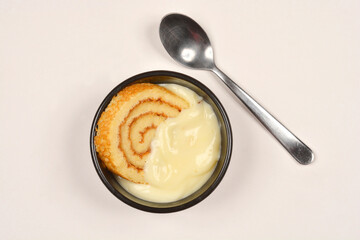 A piece of Swiss roll with cream is served in a small black bowl next to a spoon on a light surface