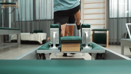 Man practicing kneeling stretch on pilates reformer with assistance to improve flexibility and strength. Trainer assist student doing lunge stretch on the reformer for enhancing balance. Habituate.