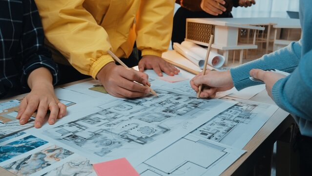 A collaborative atmosphere in an architecture office where multiple hands engage in sketching a detailed floor plan, surrounded by design materials and creative models. SACTR