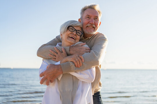 Senior mature couple hugging enjoying outdoor recreation walking on beach. Old husband wife embracing with tenderness love enjoying sweet bonding. Grandmother grandfather together. Family moment love - Powered by Adobe