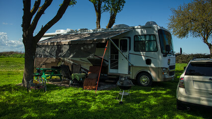 Rv motorhome parked at campsite with bike and pit and chairs under trees and blue sky