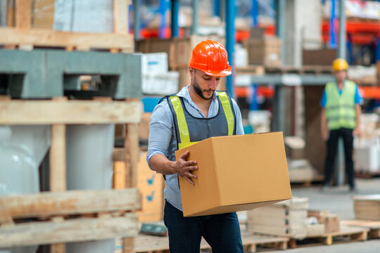 Caucasian man storage worker in uniform and hard hat carries box in hands. Images of the warehouse worker carrying cardboard boxes of the delivery industry, safely in uniform helmets for protection
