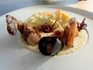 A fine dining seafood dish featuring shrimp, mussels, and squid over a creamy sauce with whipped topping and thinly sliced fruit.