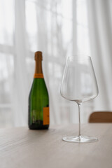 Fresh green champagne bottle with large champagne and wine glass on wooden table in the early morning with white curtains in the background. Minimalist room interior