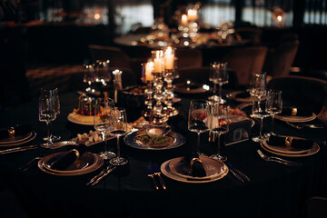 Elegant indoor dinner party table setting with a black tablecloth, gold-rimmed plates, dark napkins, and a silver candelabra with lit candles