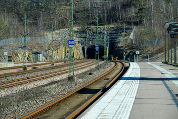 Train tracks curve toward a dark tunnel at a quiet station. Signals indicate train movement,...