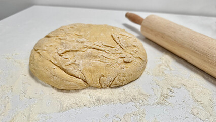 Fresh Dough Ready for Baking with a Rolling Pin and Flour Dusting. Raw dough ball on a floured surface, accompanied by a wooden rolling pin, set for baking preparations. Captures the essence of baking