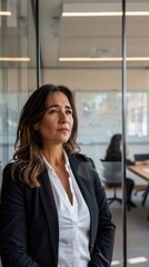 Confident middle-aged Latina businesswoman standing in front of glass wall in modern office, wearing professional business attire, calm and serious expression, corporate environment
