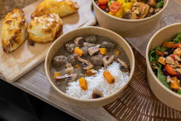 Sustainable bowl filled with white rice and meatballs in creamy mushroom sauce, garnished with sauteed mushrooms and squash. Fresh and convenient meal-to-go, plastic-free container. Wooden background.