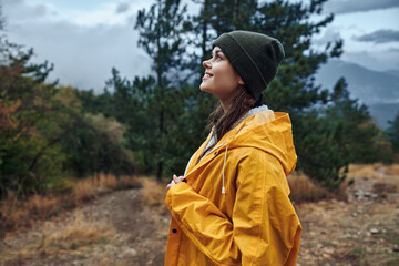 A woman in a yellow raincoat standing on a hill overlooking the majestic mountains in the...