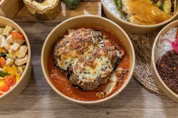 A bowl of oven-baked stuffed eggplant topped with melted cheese over rich tomato sauce. Served in an eco-friendly container. Horizontal on wooden surface. Artisanal takeout dish, compostable food box.