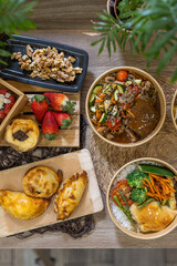 Flatlay of empanadas, strawberries, nuts, dessert, and healthy bowls with vegetables and meat. Presented on rustic surfaces and wooden boards and greenery. Vertical image, copy-space.