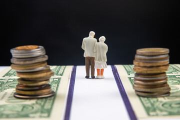 Two miniature elderly figurines stand together on a white strip between stacks of coins and dollar bills, symbolizing financial journey or retirement planning against a dark background.