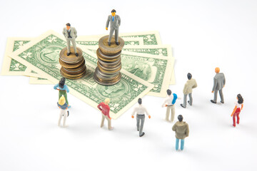 Artistic shot of small figurines in a row, juxtaposed with stacks of coins and U.S. currency,...