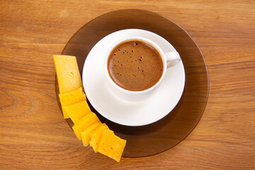 minimalist setup showcasing a steaming espresso in a white cup on a saucer, paired with neatly arranged yellow cheese slices, set against a rustic wooden background.