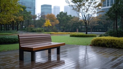 Fototapeta premium A park bench, made of dark brown wood with a metal frame, sits on a wet wooden deck. The background shows a city park with lush greenery and modern buildings. The image is high-quality, with natural