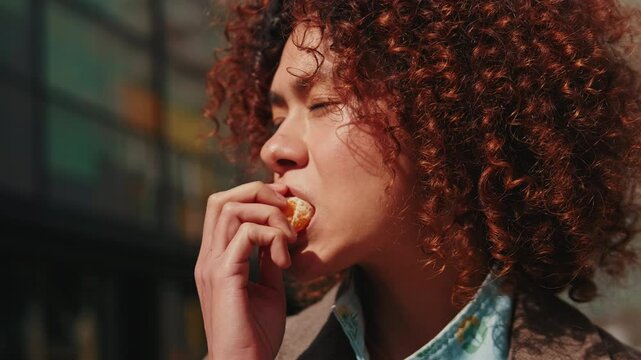 A young woman with curly red hair enjoys a fresh and juicy mandarin orange on a sunny day outdoors.