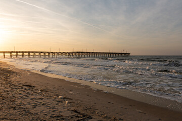 Waves crash along the shore as Oak Island Pier extends into the Atlantic Ocean under a glowing sunrise sky in North Carolina.