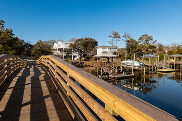 A scenic wooden footbridge with railings leads through a tranquil coastal neighborhood with elevated houses, lush trees, and a calm waterway under a clear blue sky.