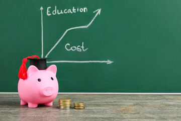 Piggy bank with graduation hat and coins on table near green blackboard. Student loan concept