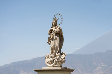 Obraz premium Photograph of a sculpture of a religious woman with angels below, outdoors on a sunny day and with mountains in the background