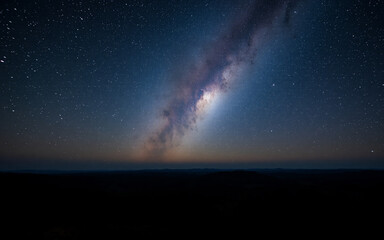 Fototapeta premium A breathtaking view of the night sky showcases the bright milky way stretching across the dark expanse above a shadowy landscape. Countless stars twinkle against the deep blue gradient.