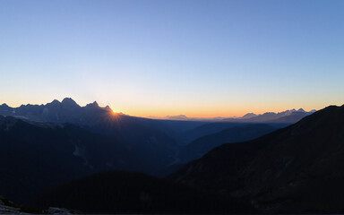 Fototapeta premium A scenic vista of mountain peaks silhouetted against the bright sunrise. The sky transitions from dark blue to orange as the sun illuminates the horizon.