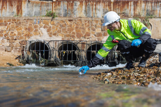 A safety-clad environmental worker collects a water sample from a rocky shoreline near an industrial drainage outlet, using scientific equipment to test for pollutants or contamination.