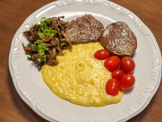Delicious homemade meal of grilled beef steak served with creamy polenta, sautéed mushrooms with scallions, and fresh cherry tomatoes on a white plate over a wooden table.