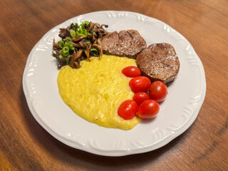 Delicious homemade meal of grilled beef steak served with creamy polenta, sautéed mushrooms with scallions, and fresh cherry tomatoes on a white plate over a wooden table.