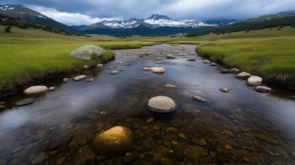 Naklejka premium Crystal clear mountain stream winding through a grassy valley, reflecting the surrounding peaks. A tranquil alpine scene under a dramatic sky