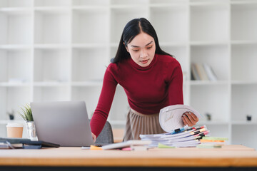 Focused woman in red sweater and beige skirt working at desk with laptop, reviewing documents in...