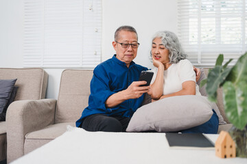 Elderly couple sitting on sofa, smiling and looking at smartphone together in cozy living room.