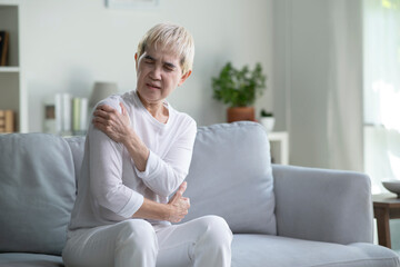 Fototapeta premium Elderly Asian woman at home with shoulder pain, Senior lady feeling muscles pain attack sitting on sofa in living room.