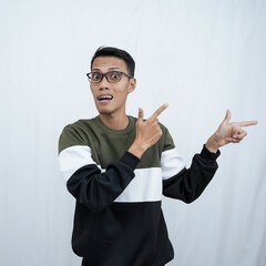 A young Asian man with glasses wearing a sweater with a shocked, surprised expression while pointing to the side of the empty space. Isolated on a white background.