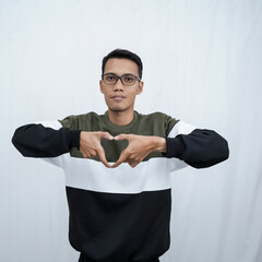 A young Asian man with glasses wearing a sweater smiles while making a love sign with his hands. Isolated on white background.