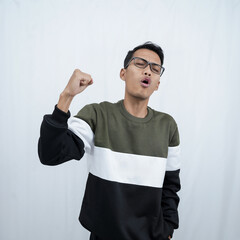 A young Asian man with glasses wearing a sweater with a happy, excited, triumphant expression while raising and clenching his fist. Isolated on white background.