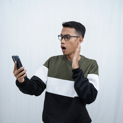 A young Asian man with glasses wearing a sweater with a surprised happy expression getting a surprise while looking at a smartphone. Isolated on a white background.