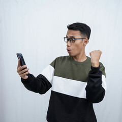 A young Asian man with glasses wearing a sweater with a surprised happy expression getting a surprise while looking at a smartphone. Isolated on a white background.
