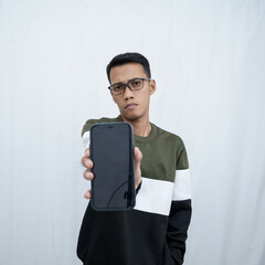 A young Asian man with glasses wearing a sweater points to a smartphone screen. Isolated on a white background.
