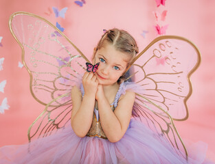 Girl in fairy wings holds pink butterfly near face on pink backdrop